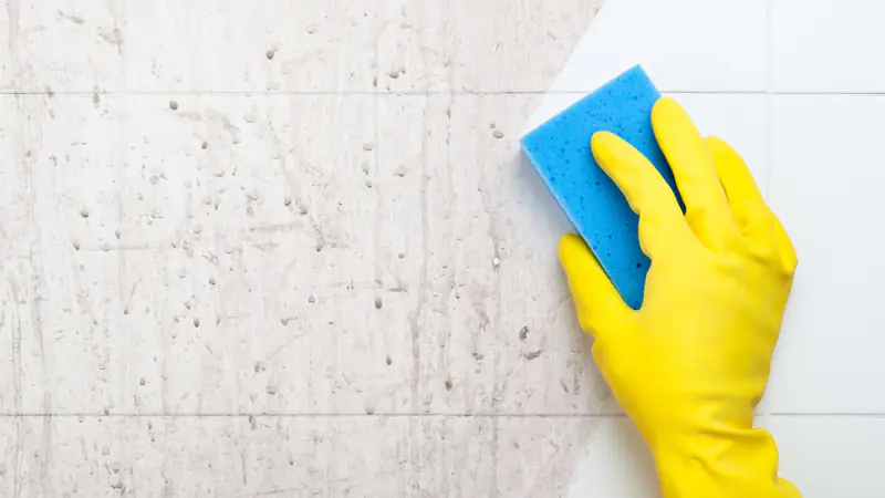 Hand im gelben Handschuh reinigt Wandfliesen mit einem blauen Schwamm